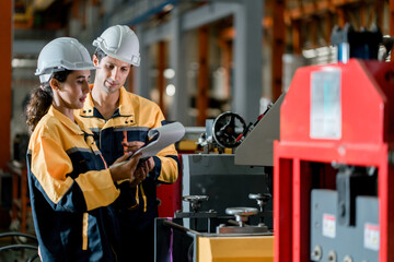 Two professional engineer,worker,technician use clipboard discuss work, walk in steel metal manufacture factory plant industry. Black African American man and woman wear hard hat check quality machine