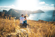 © Michael - Back view of a tourist couple sitting on a hillside, overlooking the coastal landscape of Madeira Island in the Atlantic Ocean in the morning. São Lourenço, Madeira Island, Portugal, Europe.