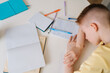 © dikushin - Close up top view of exhausted pupil boy sitting at home or classroom lying on desk filled with books training material. Frustrated little child boy sleeping while doing homework with head on table