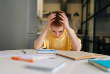 Portrait Of Exhausted Pupil Boy Tired From Studying Holding Head Head With Hands Sitting At Desk With Paper Copybook, Looking Down. Frustrated Child Schoolboy Doing Homework At Home.