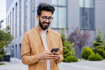 Successful Indian young businessman outside office building walking in daytime, man holding phone in hands, businessman dialing, browsing online pages, programmer engineer in glasses.