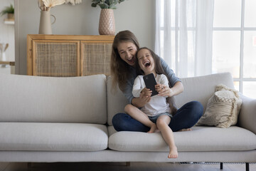 Wall Mural - Positive excited young mom holding cheerful cute little kid in arms on lap, taking selfie picture on mobile phone, relaxing on comfortable couch, laughing at smartphone screen