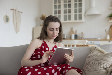 Focused beautiful young adult girl using modern technology, Internet application on tablet, shopping online, buying trendy clothes on webstores, resting on sofa with digital gadget