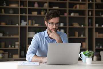 Wall Mural - Focused young businessman working at laptop computer. Freelancer using modern Internet technology for job, thinking on freelance business project, touching chin, looking at screen