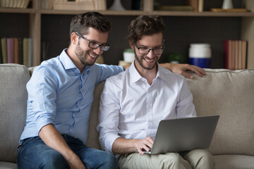 Wall Mural - Cheerful young twin brother men in glasses using laptop for Internet job communication, working at home together, brainstorming on freelance business project