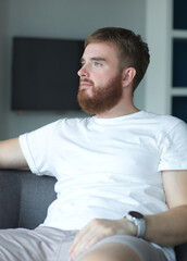 Wall Mural - Portrait young european beard pensive thoughtful relaxed lonely guy, man is dreaming, thinking, relax at home in living room at comfortable sofa, couch with his hands behind head 