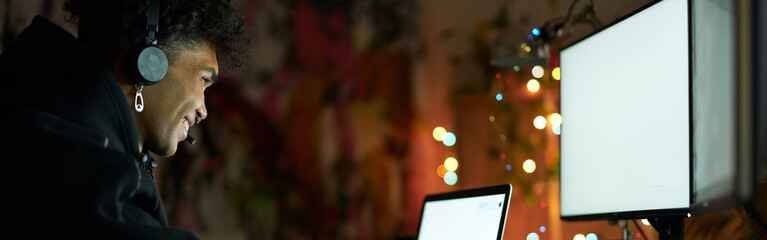 night shift. smiling young guy wearing headset sitting at the table in front of computer monitors an