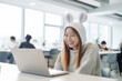 © Surachetsh - A business-focused Asian girl works on her laptop in a modern co-working space, while wearing a fun rabbit knit hat that adds a touch of unique style. generative AI.