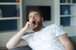 © Евгений Шемякин - young man at home sitting on the sofa tired yawning, sleepless,bored in the living room at home interior