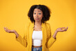 Doubtful black lady shrugging shoulders and spreading arms, unsure hesitant woman standing over yellow studio background