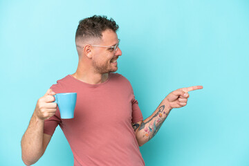 Wall Mural - Young caucasian man holding cup of coffee isolated on blue background pointing to the side to present a product