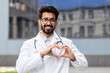 © Liubomir - Portrait of friendly doctor, Hindu man in white medical coat smiling and looking at camera, showing heart with hands gesture of kindness.