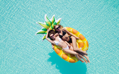 Wall Mural - Top view of two girls relaxing on inflatable big pineapple in the pool. Aerial view of ladies relaxing on the floating mattress.