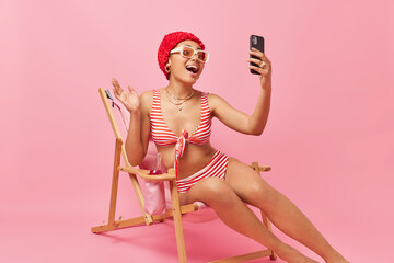 People summer vacation and resort concept. Positive smiling young slim woman poses on deck chair uses mobile phone broadcasting livestream from vacation wears bikini smiles gladfully has video call