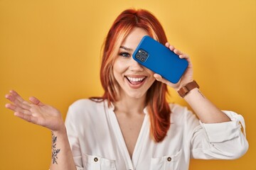 Canvas Print - Young caucasian woman covering eye with smartphone celebrating achievement with happy smile and winner expression with raised hand