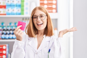 Poster - Young redhead woman working at pharmacy drugstore holding condom celebrating victory with happy smile and winner expression with raised hands