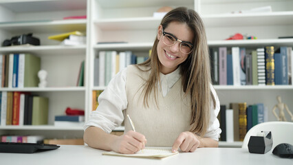 Young beautiful hispanic woman student writing on notebook at the library