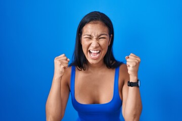 Poster - Hispanic woman standing over blue background angry and mad raising fists frustrated and furious while shouting with anger. rage and aggressive concept.