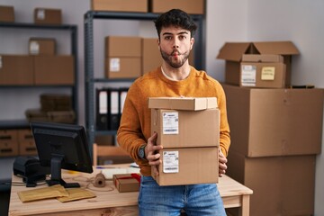 Wall Mural - Hispanic man with beard working at small business ecommerce holding packages making fish face with mouth and squinting eyes, crazy and comical.