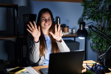 Wall Mural - Young brunette woman working at the office at night showing and pointing up with fingers number eight while smiling confident and happy.