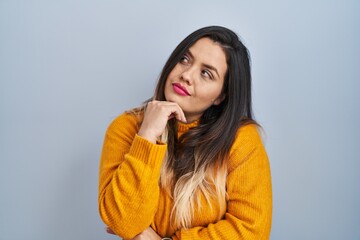 Wall Mural - Young hispanic woman standing over isolated background with hand on chin thinking about question, pensive expression. smiling with thoughtful face. doubt concept.