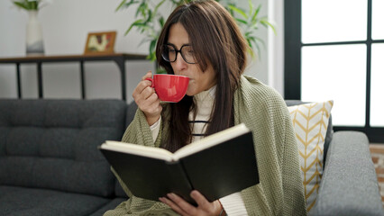 Poster - Young beautiful hispanic woman reading book and drinking coffee sitting on sofa at home