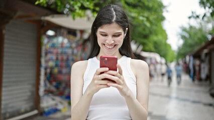 Wall Mural - Young beautiful hispanic woman using smartphone smiling at street market