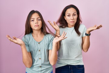 Canvas Print - Young mother and daughter standing over pink background clueless and confused expression with arms and hands raised. doubt concept.