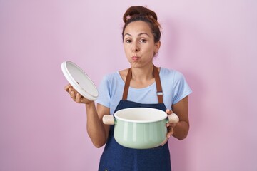 Canvas Print - Brunette woman wearing apron holding cooking pot puffing cheeks with funny face. mouth inflated with air, catching air.