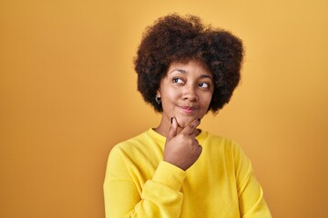 Poster - Young african american woman standing over yellow background looking confident at the camera smiling with crossed arms and hand raised on chin. thinking positive.