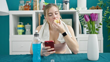 Young Blonde Woman Wearing Sportswear Eating Apple Using Smartphone At Dinning Room