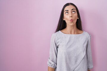 Wall Mural - Young brunette woman standing over pink background making fish face with lips, crazy and comical gesture. funny expression.