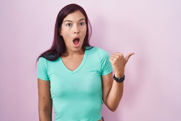 Wall Mural - Young hispanic woman standing over pink background surprised pointing with hand finger to the side, open mouth amazed expression.