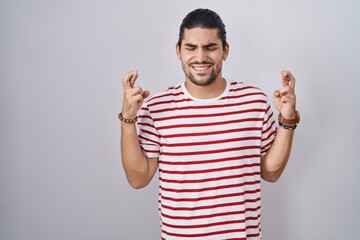 Sticker - Hispanic man with long hair standing over isolated background gesturing finger crossed smiling with hope and eyes closed. luck and superstitious concept.