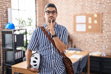 Canvas Print - Hispanic man with long hair working at the office holding bike helmet thinking concentrated about doubt with finger on chin and looking up wondering