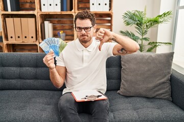 Sticker - Young hispanic man working on couple therapy at consultation office holding chilean pesos with angry face, negative sign showing dislike with thumbs down, rejection concept