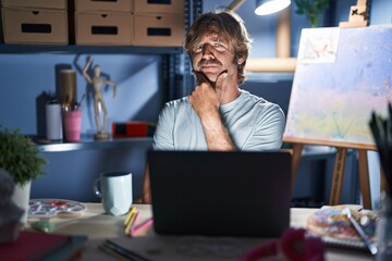 Poster - Middle age man sitting at art studio with laptop at night looking confident at the camera with smile with crossed arms and hand raised on chin. thinking positive.