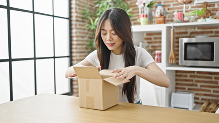 Sticker - Young chinese woman unpacking cardboard box sitting on table with serious expression at dinning room