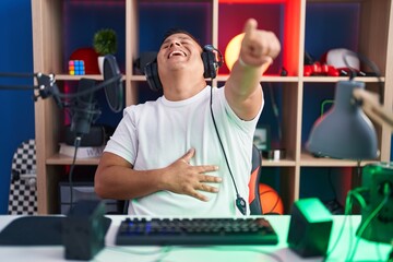 Poster - Young hispanic man playing video games laughing at you, pointing finger to the camera with hand over body, shame expression