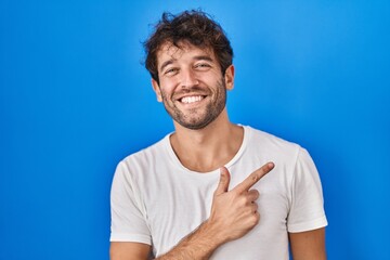 Poster - Hispanic young man standing over blue background cheerful with a smile on face pointing with hand and finger up to the side with happy and natural expression