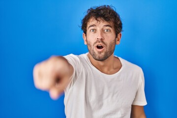 Poster - Hispanic young man standing over blue background pointing with finger surprised ahead, open mouth amazed expression, something on the front