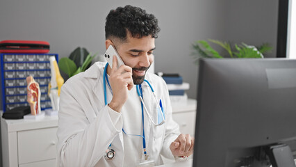 Wall Mural - Young hispanic man doctor talking on smartphone using computer at clinic