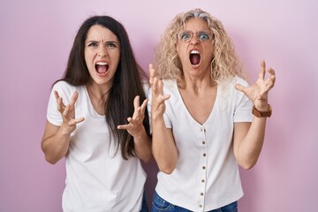 Sticker - Mother and daughter standing together over pink background crazy and mad shouting and yelling with aggressive expression and arms raised. frustration concept.