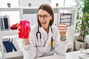 Sticker - Young caucasian dentist woman holding denture and mouthwash angry and mad screaming frustrated and furious, shouting with anger. rage and aggressive concept.