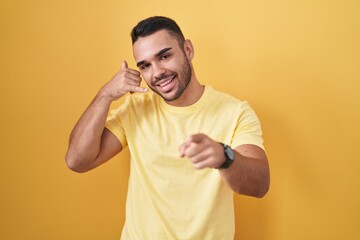 Canvas Print - Young hispanic man standing over yellow background smiling doing talking on the telephone gesture and pointing to you. call me.