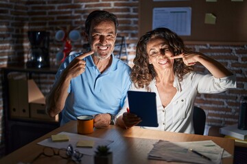 Canvas Print - Middle age hispanic couple using touchpad sitting on the table at night smiling cheerful showing and pointing with fingers teeth and mouth. dental health concept.