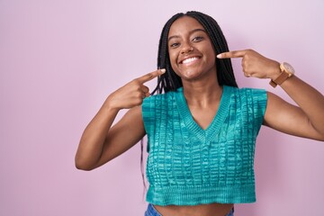 Sticker - Young african american with braids standing over pink background smiling cheerful showing and pointing with fingers teeth and mouth. dental health concept.