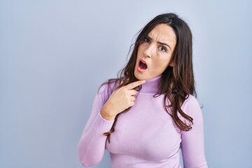 Poster - Young brunette woman standing over blue background in shock face, looking skeptical and sarcastic, surprised with open mouth