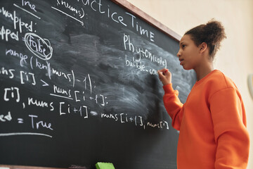 Side view portrait of black teenage girl writing on blackboard in school classroom, copy space