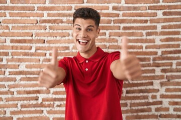 Sticker - Young hispanic man standing over bricks wall approving doing positive gesture with hand, thumbs up smiling and happy for success. winner gesture.